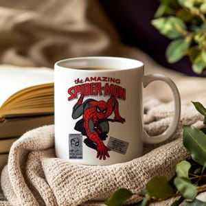 May include: White ceramic mug featuring a comic book illustration of Spider-Man. The mug displays the text "The Amazing Spider-Man" in red and white, with a detailed image of the superhero in action. The mug is set on a neutral-toned blanket.
