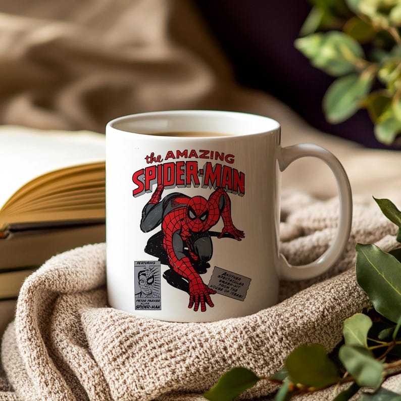 May include: White ceramic mug featuring a comic book illustration of Spider-Man. The mug displays the text "The Amazing Spider-Man" in red and white, with a detailed image of the superhero in action. The mug is set on a neutral-toned blanket.