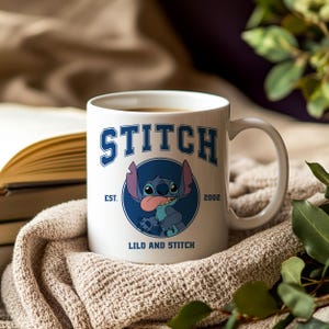 Puede incluir: Taza de cer&aacute;mica blanca con la palabra "STITCH" en azul y un dibujo del personaje Stitch. La taza tambi&eacute;n incluye el texto "EST. 2002" y "LILO AND STITCH". La taza est&aacute; llena de una bebida caliente.