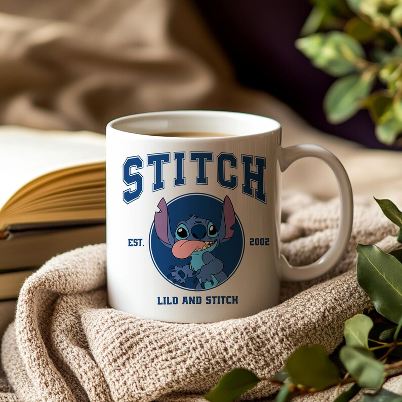 Puede incluir: Taza de cer&aacute;mica blanca con la palabra "STITCH" en azul y un dibujo del personaje Stitch. La taza tambi&eacute;n incluye el texto "EST. 2002" y "LILO AND STITCH". La taza est&aacute; llena de una bebida caliente.
