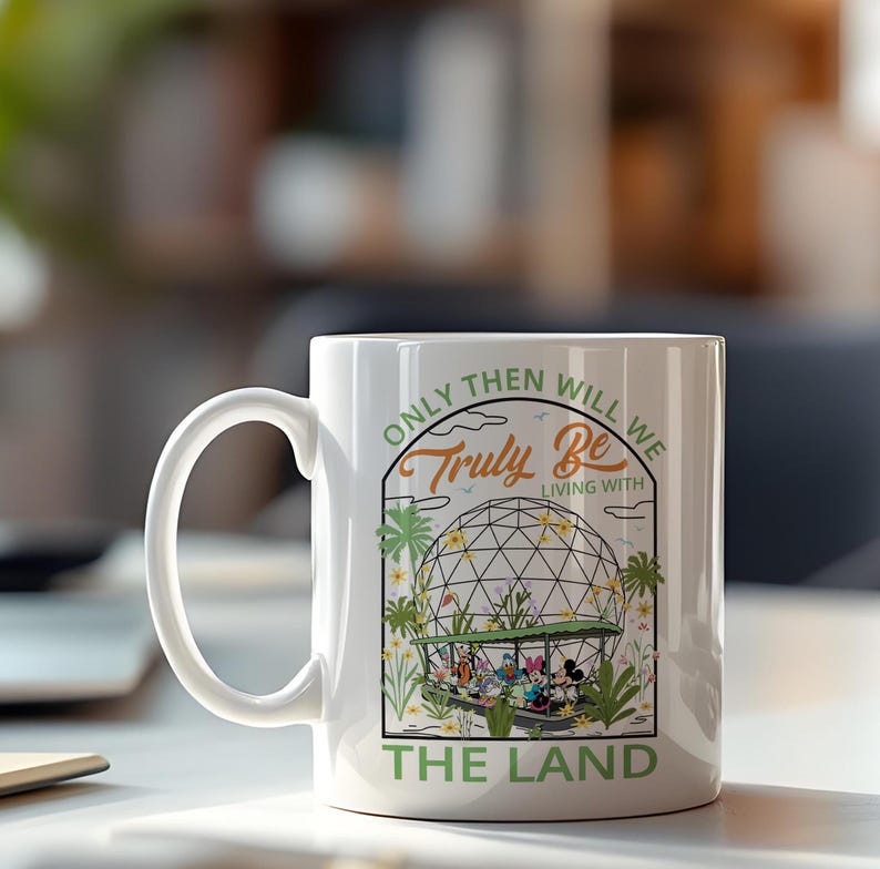 May include: White ceramic mug featuring a colorful graphic of Disney characters in a greenhouse setting, with the text "ONLY THEN WILL WE TRULY BE LIVING WITH THE LAND". The mug has a curved handle.