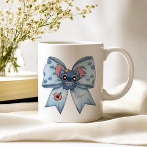 Pu&ograve; includere: Tazza in ceramica bianca con un personaggio dei cartoni animati blu con grandi orecchie e una corona, su un fiocco azzurro con pois bianchi. La parola "Ohana" &egrave; scritta sul fiocco. C'&egrave; anche un piccolo fiore rosso.