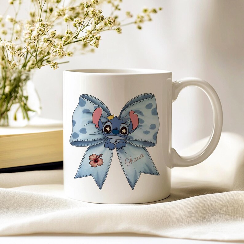 Pu&ograve; includere: Tazza in ceramica bianca con un personaggio dei cartoni animati blu con grandi orecchie e una corona, su un fiocco azzurro con pois bianchi. La parola "Ohana" &egrave; scritta sul fiocco. C'&egrave; anche un piccolo fiore rosso.