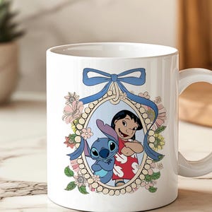May include: White ceramic mug featuring a colorful illustration of Lilo and Stitch. The design includes a blue bow, floral accents, and a framed image of the characters. The mug has a curved handle for easy gripping.