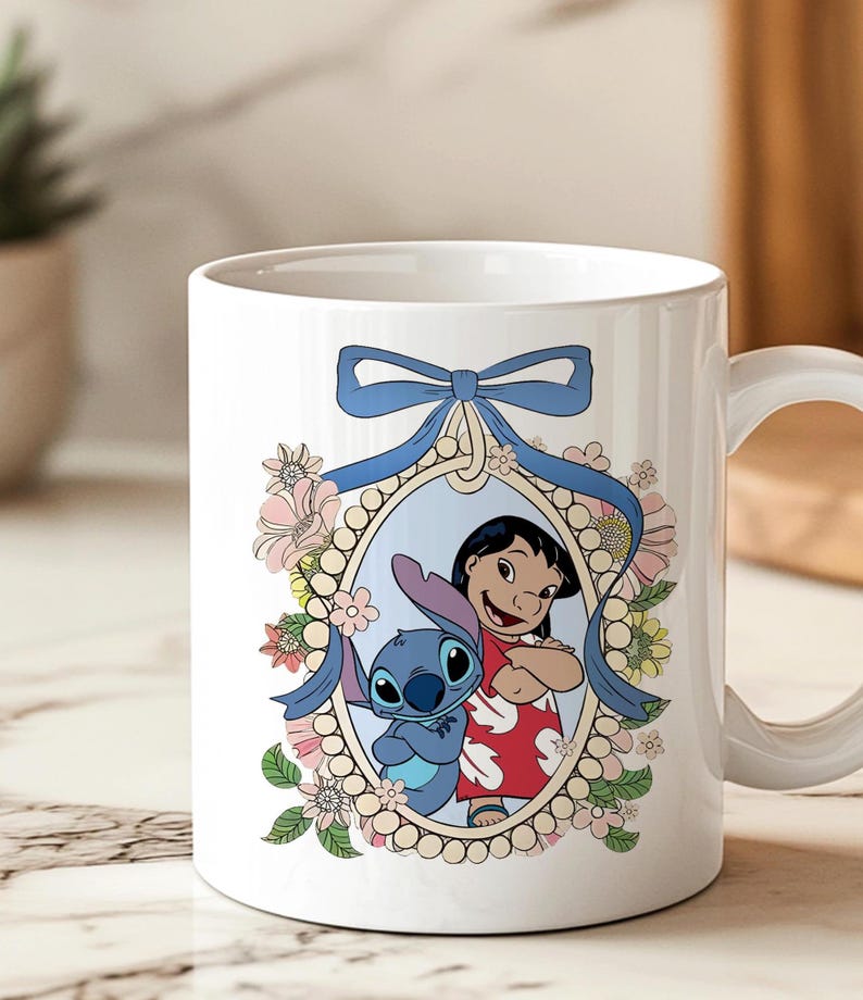 May include: White ceramic mug featuring a colorful illustration of Lilo and Stitch. The design includes a blue bow, floral accents, and a framed image of the characters. The mug has a curved handle for easy gripping.