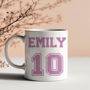 May include: White ceramic mug with the name "EMILY" in a pink, distressed font above the number "10" in a pink, outlined font. The mug has a white handle and is set against a neutral background.