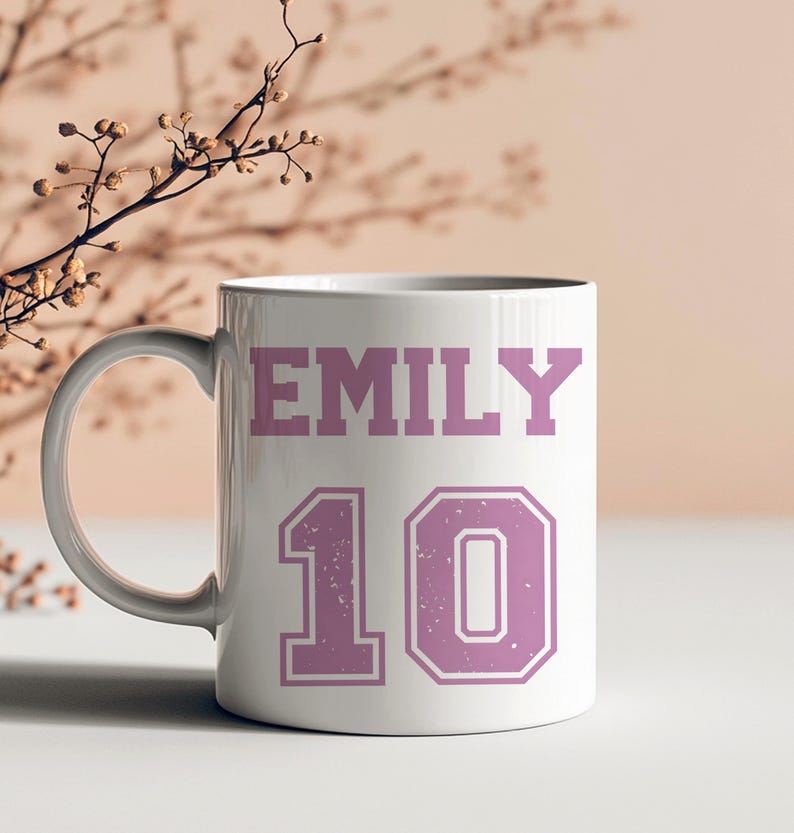 May include: White ceramic mug with the name "EMILY" in a pink, distressed font above the number "10" in a pink, outlined font. The mug has a white handle and is set against a neutral background.