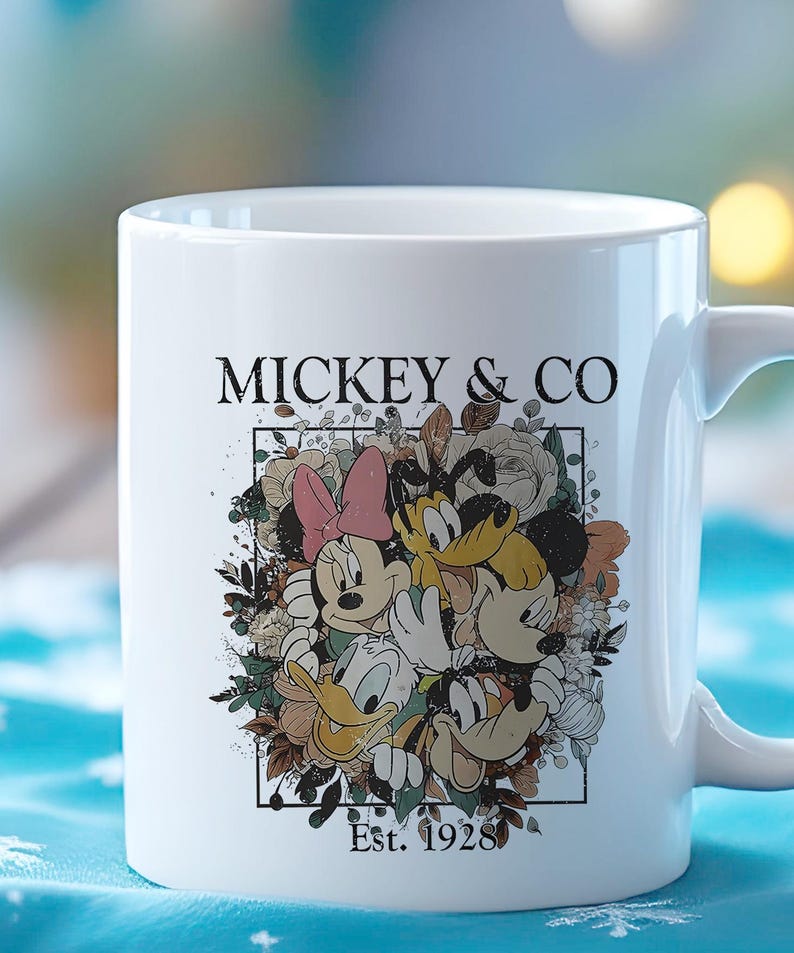 May include: White ceramic mug with a floral design featuring Mickey Mouse, Minnie Mouse, Donald Duck, and Goofy. The text "Mickey & Co. Est. 1928" is printed on the mug.
