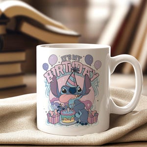 May include: White ceramic mug featuring a cartoon character with the text "IT'S MY BIRTHDAY". The character is blue with large ears and is wearing a party hat. The mug also has a birthday cake and presents.