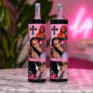 May include: Two cylindrical tumblers with black lids and clear glitter straws. The exterior features a pink collage of photographic portraits, a black cross icon, cherry graphics, and the text THEREALIYANNA printed in a stylized white font.