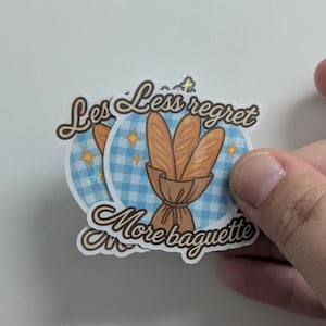 May include: A set of stickers with a blue gingham background and the text "Less regret, More baguette." The stickers feature illustrations of baguettes in a brown paper bag, with gold star accents.