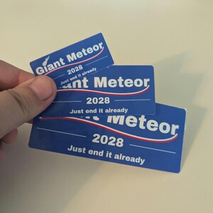 May include: Blue cards with the text "Giant Meteor 2028 Just end it already" in white and red. The cards are held in a hand. The cards have a blue background with a white and red curved line.