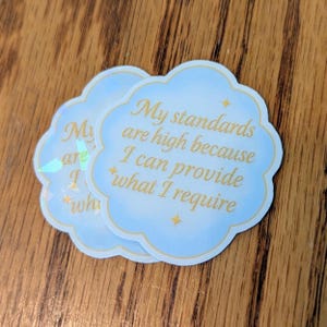 May include: Two cloud-shaped stickers with a light blue background and gold text. The text reads "My standards are high because I can provide what I require." The stickers have a holographic sheen.