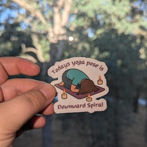 May include: A sticker with the text "Todays yoga pose is Downward Spiral" shows a cartoon figure in a yoga pose on a purple mat, with lit candles. The figure wears a teal shirt and has brown hair.