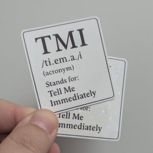 May include: Two white rectangular stickers with the acronym "TMI" in bold black letters. The stickers define "TMI" as "Tell Me Immediately". One sticker has a holographic background.