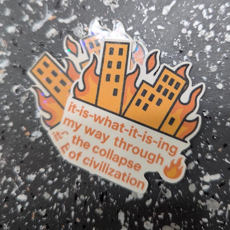 May include: A sticker featuring orange buildings engulfed in flames. The text reads "it-is-what-it-is-ing my way through the collapse of civilization." The sticker has a holographic effect.