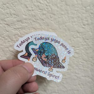 May include: A holographic sticker with the text "Todays yoga pose is downward spiral". The sticker shows a yoga pose with candles and has a glittery, iridescent finish. The sticker is a yoga-themed item.