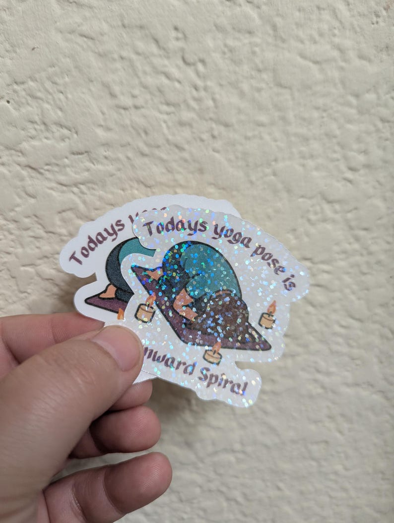 May include: A holographic sticker with the text "Todays yoga pose is downward spiral". The sticker shows a yoga pose with candles and has a glittery, iridescent finish. The sticker is a yoga-themed item.