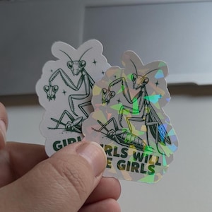 May include: Two stickers featuring a green praying mantis illustration. One sticker is holographic, and the other is white. The text "GIRLS WILL BE GIRLS" is visible on the holographic sticker. The stickers have a die-cut shape.
