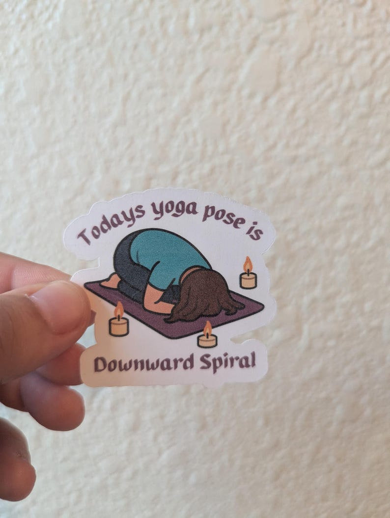 May include: A sticker with a cartoon illustration of a person in a yoga pose. The person is in a downward spiral pose on a purple mat, with candles around. The text on the sticker reads "Todays yoga pose is Downward Spiral."