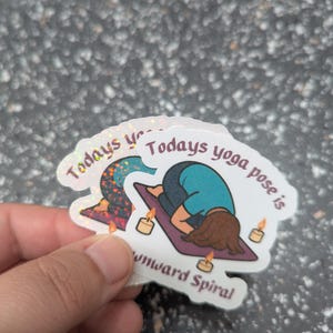 May include: Holographic stickers with the text "Todays yoga pose is downward spiral". The stickers depict a yoga pose illustration, candles, and a yoga mat.