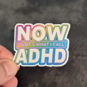 May include: A sticker with the text "NOW THAT'S WHAT I CALL ADHD" in a colorful, outlined font. The sticker has a white background with a rainbow gradient effect and a blue outline.