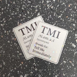 May include: Two white stickers with the acronym "TMI" in large, dark letters. The stickers also include the pronunciation and the phrase "Stands for: Tell Me Immediately."