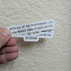 May include: Two white stickers with holographic glitter accents. The top sticker reads "WITH ALL OF THE OVERTHINKING I DO, YOU WOULD THINK I'D MAKE BETTER DECISIONS." The bottom sticker reads "DECISIONS." The stickers are held by a hand.