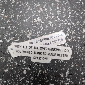May include: Two white stickers with black text on a speckled background. The stickers read: "THE OVERTHINKING I DO, MAKE BETTER" and "WITH ALL OF THE OVERTHINKING I DO, YOU WOULD THINK I'D MAKE BETTER DECISIONS."