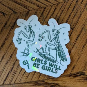 May include: Two stickers with a praying mantis illustration. The stickers have a white background with a green mantis design and the text "Girls Will Be Girls". The stickers have a holographic effect.