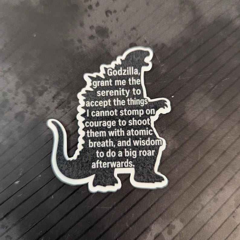 Op de afbeelding: Een zwart-witte Godzilla-vormige sticker met de tekst "Godzilla, geef me de sereniteit om de dingen te accepteren die ik niet kan vertrappen, de moed om ze met atoomadem te beschieten, en de wijsheid om daarna luid te brullen."