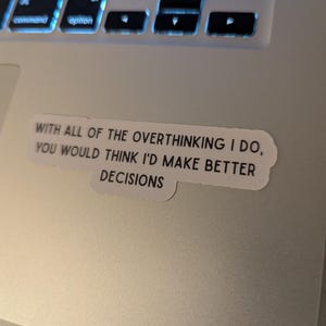 May include: A sticker with the text "WITH ALL OF THE OVERTHINKING I DO, YOU WOULD THINK I'D MAKE BETTER DECISIONS" is placed on a laptop. The laptop keyboard is visible in the background.