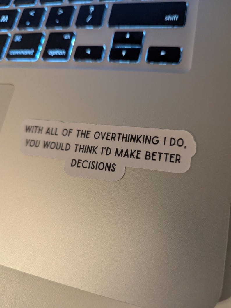 May include: A sticker with the text "WITH ALL OF THE OVERTHINKING I DO, YOU WOULD THINK I'D MAKE BETTER DECISIONS" is placed on a laptop. The laptop keyboard is visible in the background.