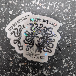 May include: A stack of holographic stickers featuring a black and white illustration of Medusa with snakes for hair. The text "MAKING MEN HARD" and "SINCE 700 BCE" are printed on the stickers.
