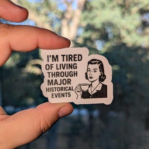 May include: A white sticker with the text "I'M TIRED OF LIVING THROUGH MAJOR HISTORICAL EVENTS" and a black and white illustration of a woman holding a teacup. The sticker is held in a hand against a blurred background.