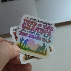 May include: A stack of holographic stickers with the text "I'M SIMULTANEOUSLY TIRED OF THIS GRANDPA AND AWARE THAT THAT'S TOO DAMN BAD". The stickers feature a desert scene with a person working and another relaxing in a rocking chair.