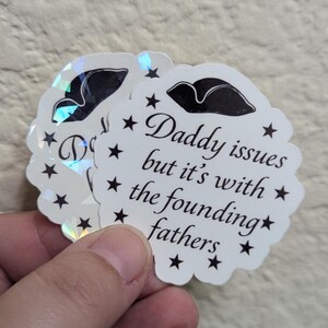 May include: A stack of white, scalloped-edge stickers with the text "Daddy issues but it's with the founding fathers" in black script. Each sticker has a black tricorn hat illustration and black star accents. The stickers have a holographic, iridescent finish.