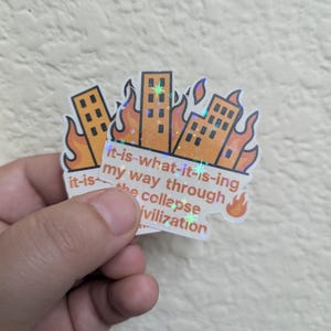 May include: A sticker with a design of buildings on fire. The sticker has the text "it-is-what-it-is-ing my way through the collapse ivilization". The sticker has a holographic effect.