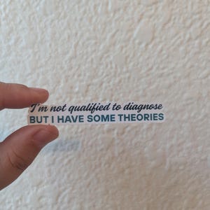 May include: A rectangular sticker with the text "I'm not qualified to diagnose BUT I HAVE SOME THEORIES" in a teal and white font. The sticker is held by a hand against a textured, off-white background.