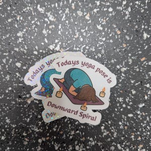 May include: A stack of holographic stickers with yoga designs. One sticker depicts a person in a downward-facing pose with the text "Todays yoga pose is Downward Spiral". The stickers have a white border and are on a speckled surface.