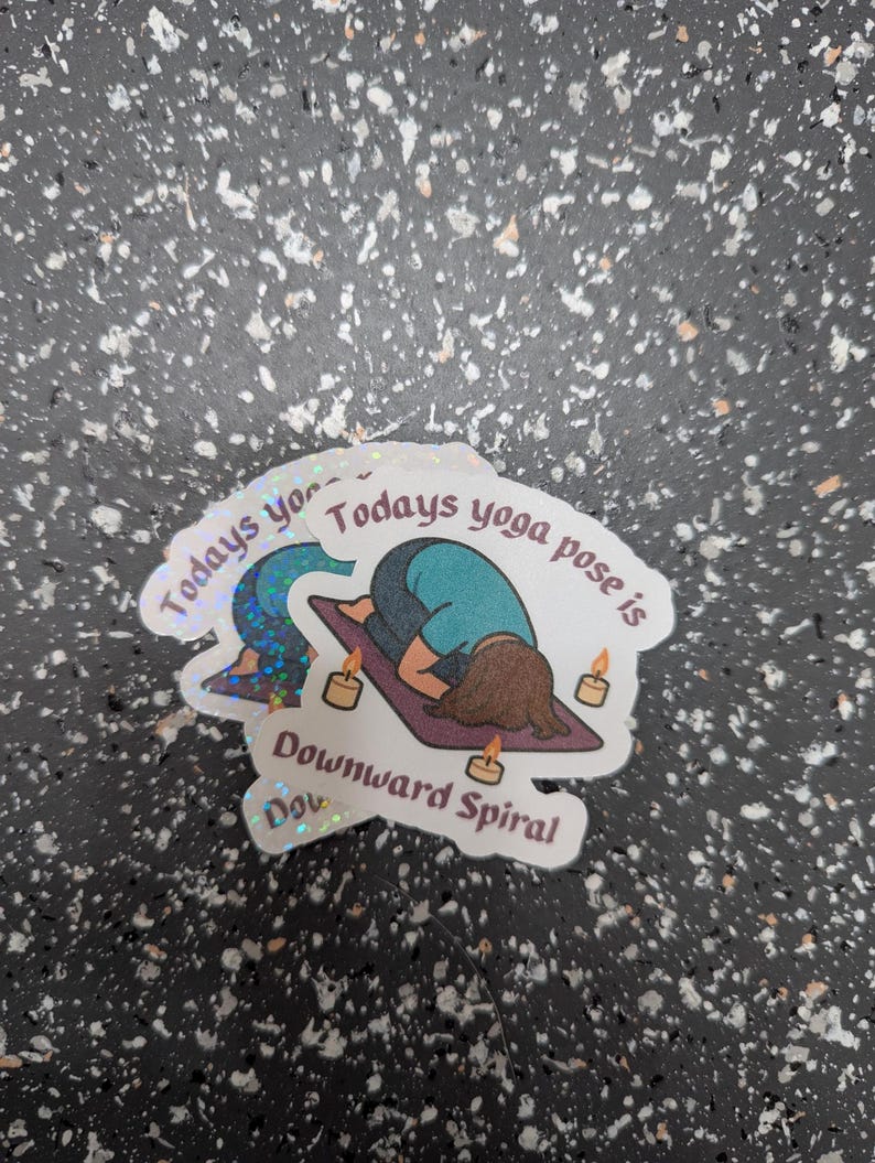May include: A stack of holographic stickers with yoga designs. One sticker depicts a person in a downward-facing pose with the text "Todays yoga pose is Downward Spiral". The stickers have a white border and are on a speckled surface.