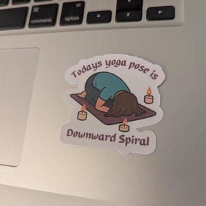 May include: A sticker with a cartoon illustration of a person in a yoga pose on a purple mat, with candles around. The text reads "Todays yoga pose is Downward Spiral" above and below the illustration.