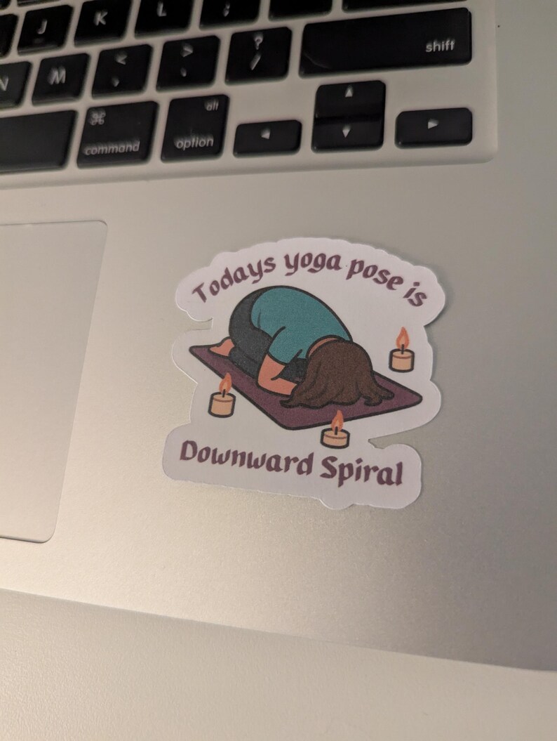 May include: A sticker with a cartoon illustration of a person in a yoga pose on a purple mat, with candles around. The text reads "Todays yoga pose is Downward Spiral" above and below the illustration.