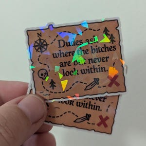 May include: Two holographic stickers with a treasure map design. The map features a compass, skull, trees, sword, and an X marking the spot. Text on the sticker reads "Dudes ask where the bitches are but never look within."