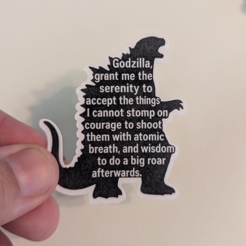 Op de afbeelding: Een zwarte sticker in de vorm van Godzilla, met de tekst: "Godzilla, geef me de sereniteit om de dingen te accepteren waar ik niet op kan stampen, de moed om ze met atoomadem te beschieten, en de wijsheid om daarna luid te brullen."