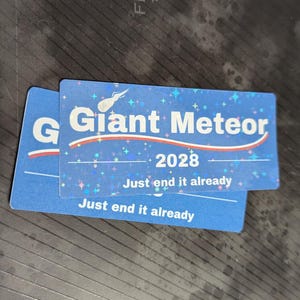 May include: Two blue rectangular stickers with the text "Giant Meteor 2028" and "Just end it already". The stickers have a comet graphic and star accents. The background is a dark, textured surface.