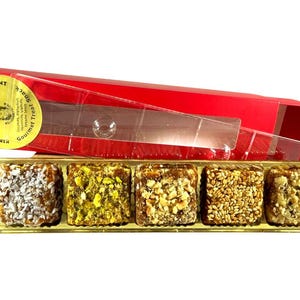 May include: A red box with a clear plastic lid contains five gourmet date treats. The dates are arranged in a gold tray and are covered in various toppings, including coconut, nuts, and seeds. The box has a label that reads "Power Dates".