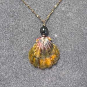May include: A gold-colored chain necklace with a pendant featuring a dark gray pearl and a colorful seashell. The seashell has yellow, orange, and green hues, with gold wire accents. The necklace is displayed on a gray surface.