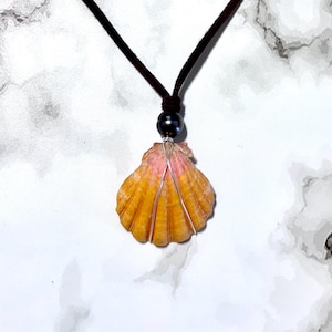 May include: A seashell pendant necklace. The pendant is a natural, orange and pink seashell, wrapped with silver wire. A black pearl bead sits above the shell, connected to a dark brown cord necklace.