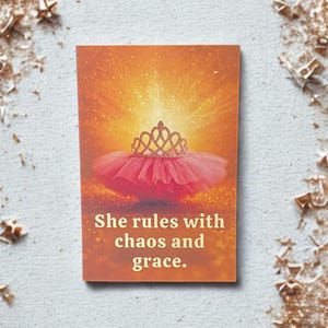 May include: A rectangular card with an orange background featuring a pink tutu and a gold crown. The text "She rules with chaos and grace" is printed in gold. The card is on a white surface with gold glitter.